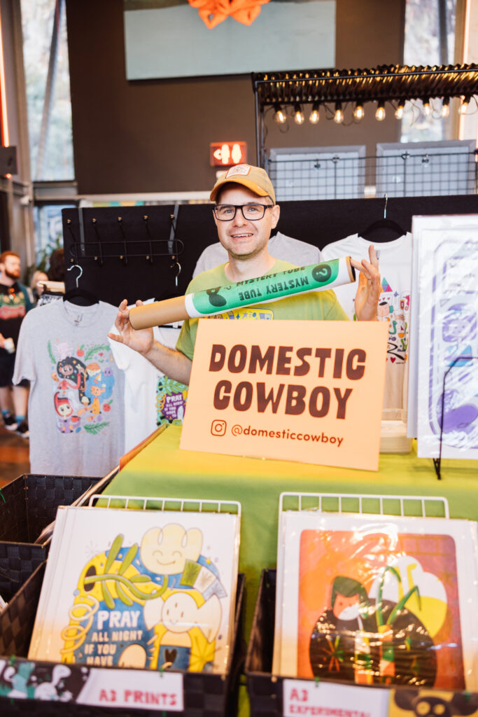 Photo of Clayton McIntosh at The Great Artist Market surrounded by his prints and tshirts. There is a sign with "Domestic Cowboy" in the middle.
